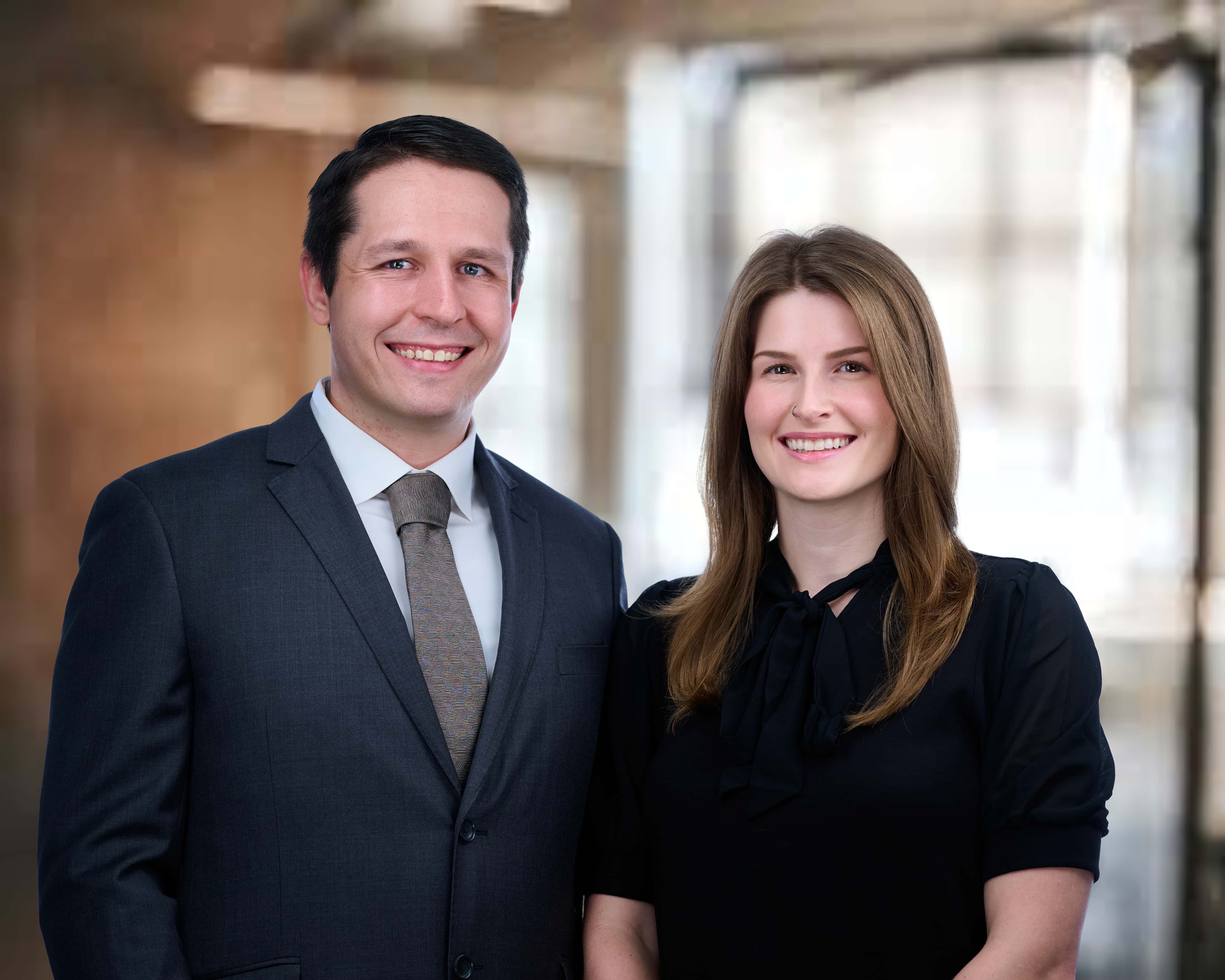 Smiling man in a dark suit and tie standing next to a smiling woman with long brown hair wearing a black blouse.