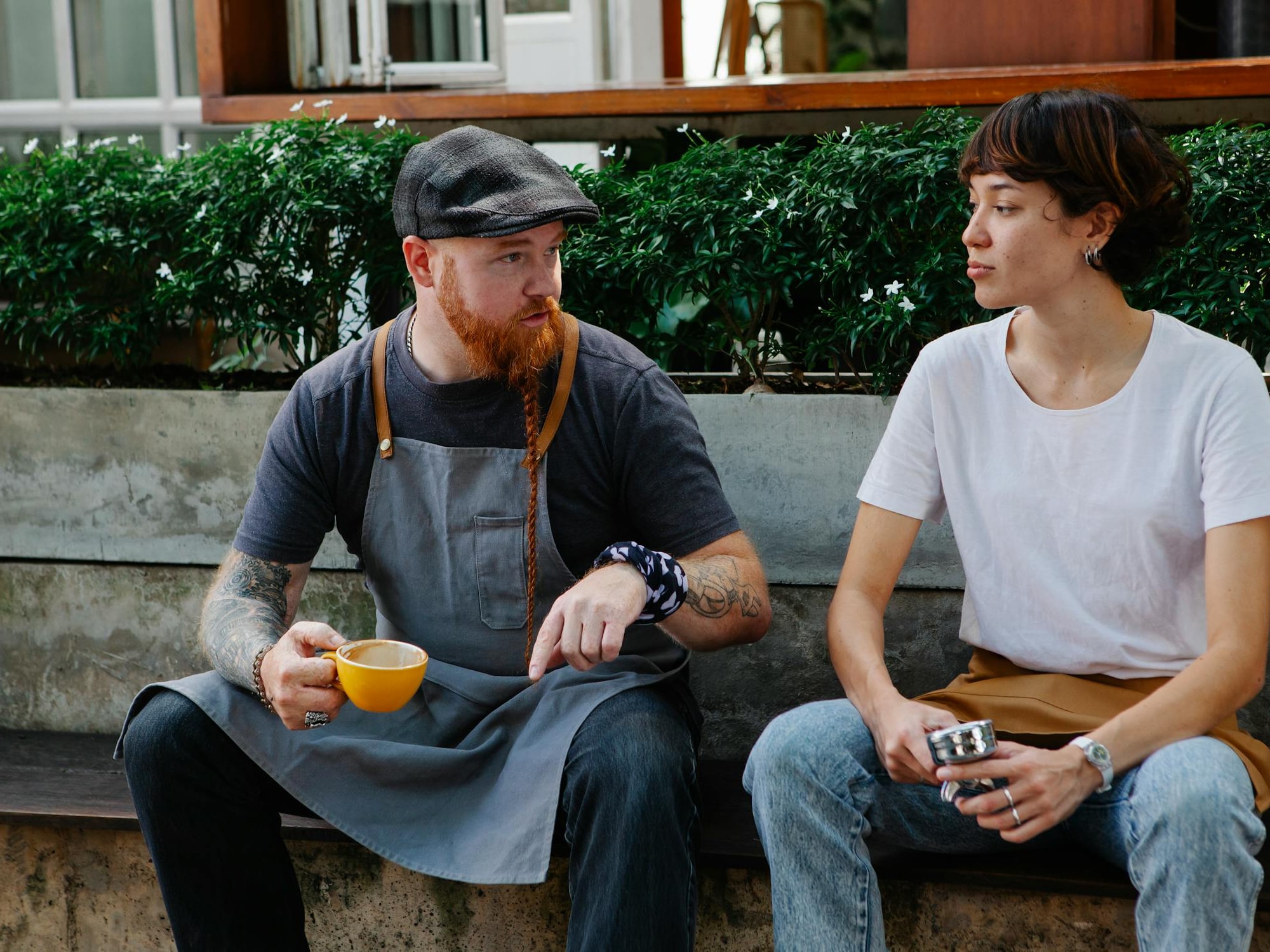 Two people sit on concrete bench, one holding yellow coffee cup, outdoors